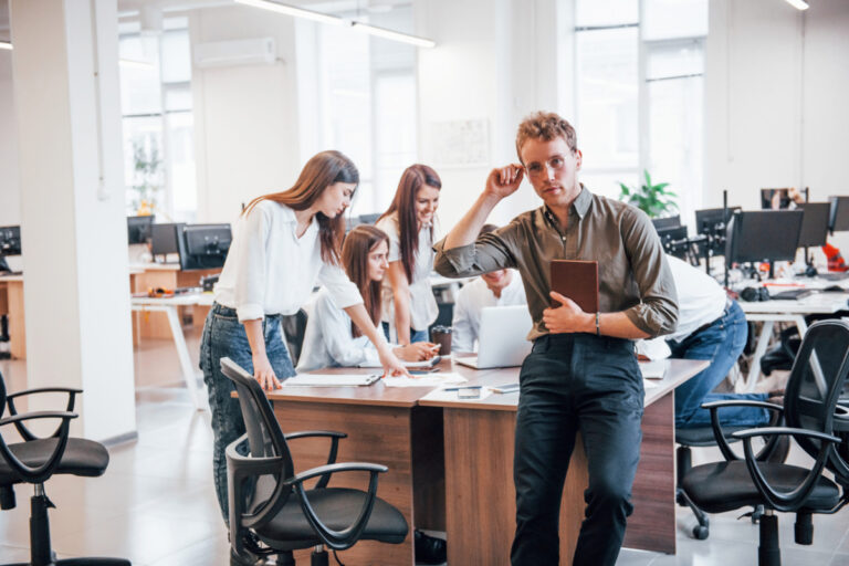 Young male employee in formal clothes standing in the office. Group of people working behind.